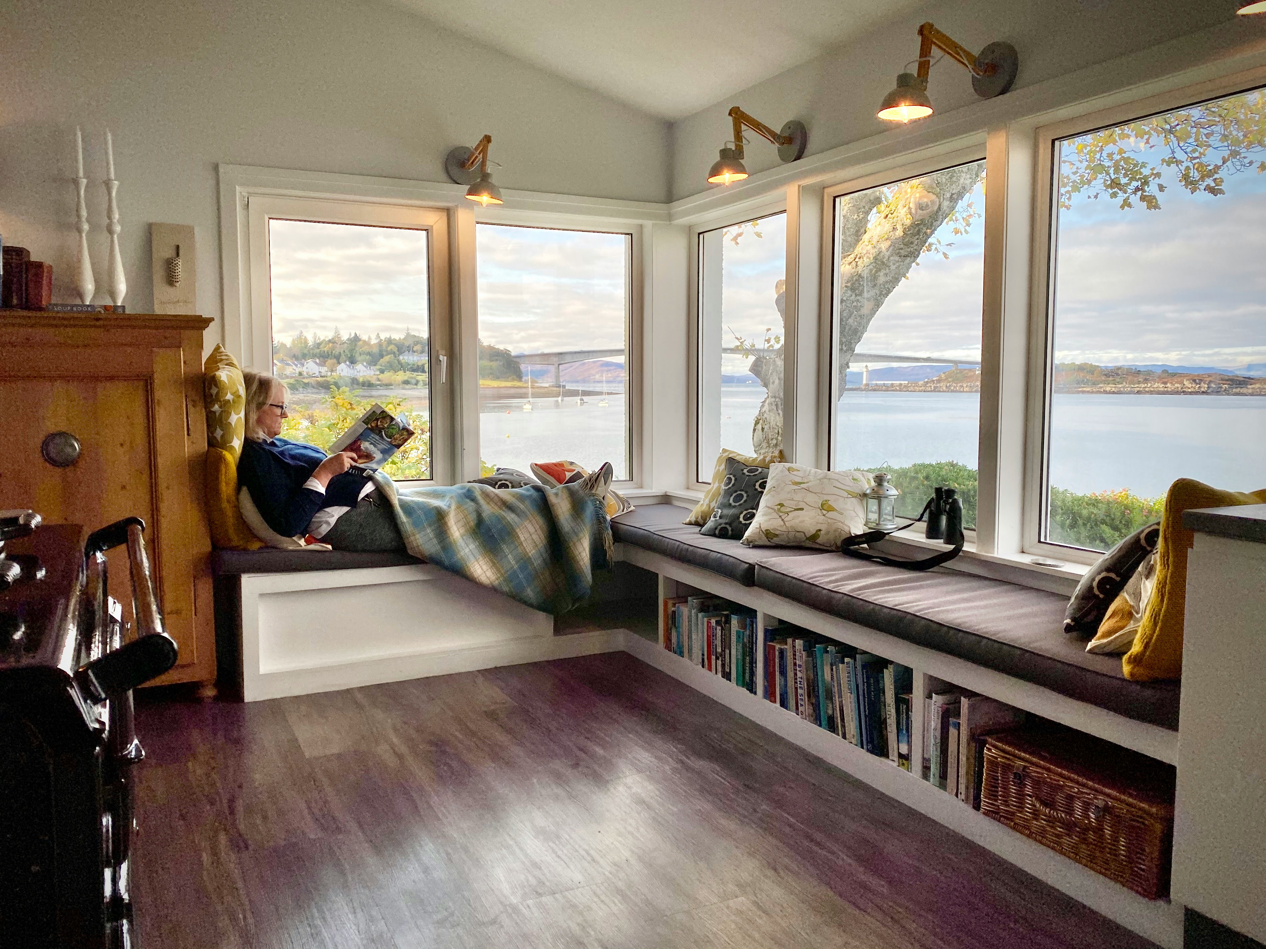A cozy reading nook with large corner windows overlooking a waterfront view. A person sits on a bench with pillows, wrapped in a blanket, reading. Outside, a bridge spans calm water under a partly cloudy sky. Photo by Richard Rawlings.