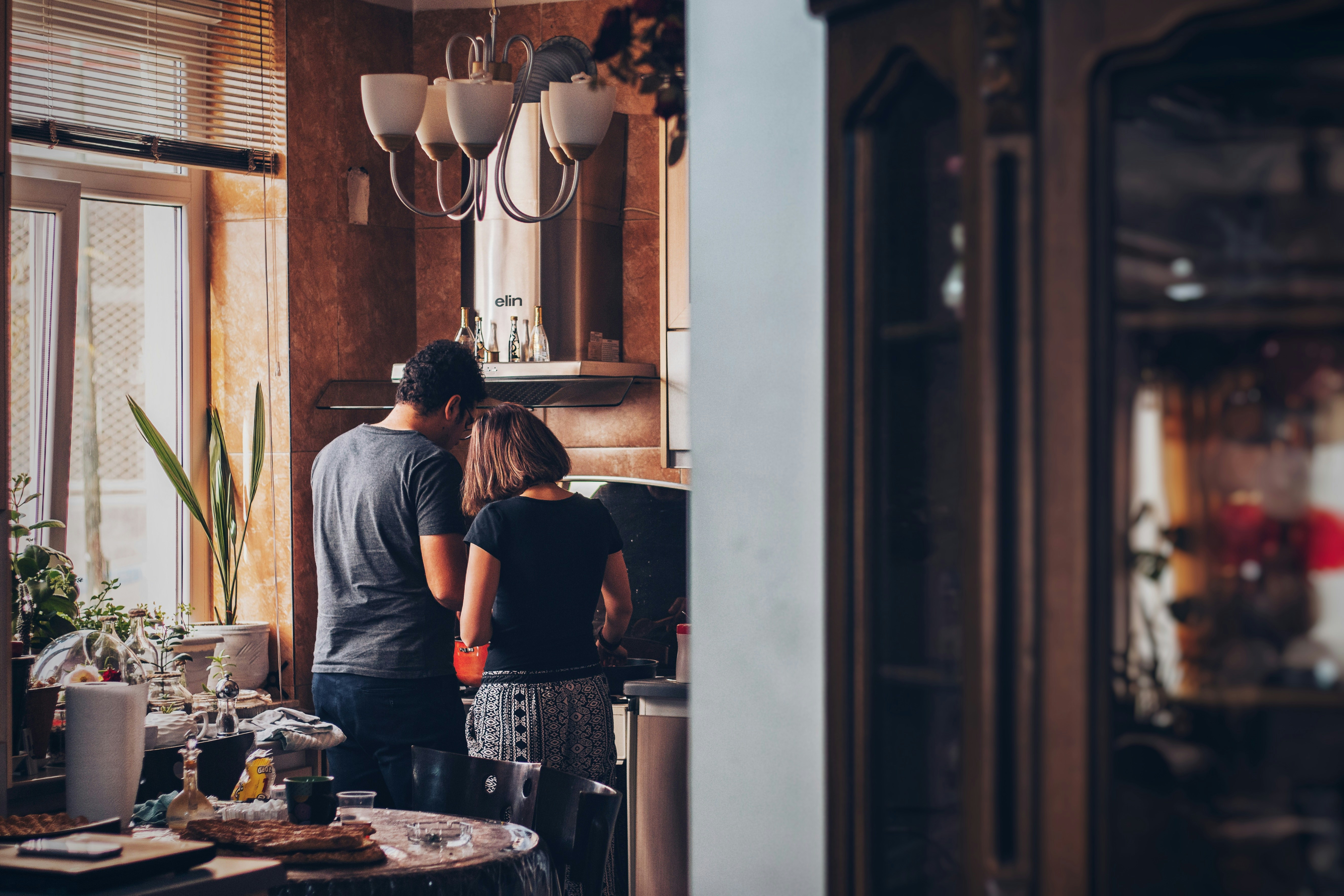  couple stands closely together in a cozy kitchen, cooking a meal. Natural light streams in from a nearby window, illuminating potted plants and various kitchen items on the counter. Photo by Soroush Karimi.