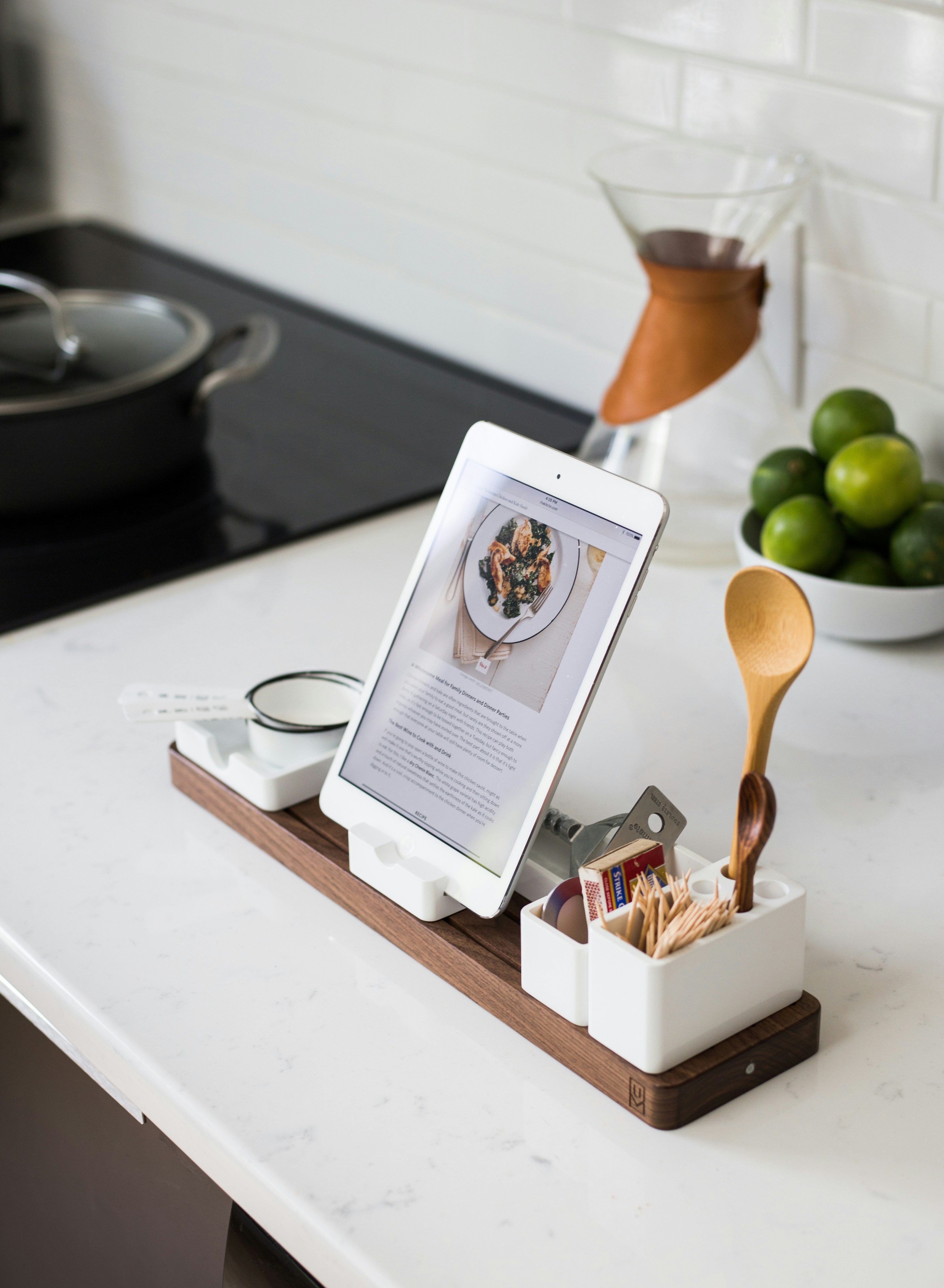 A white kitchen countertop with a tablet displaying a recipe, surrounded by an organized utensil holder, small dishes, and a wooden tray. A Chemex coffee maker and a bowl of limes add a touch of color. Photo by  Jeff Sheldon.
